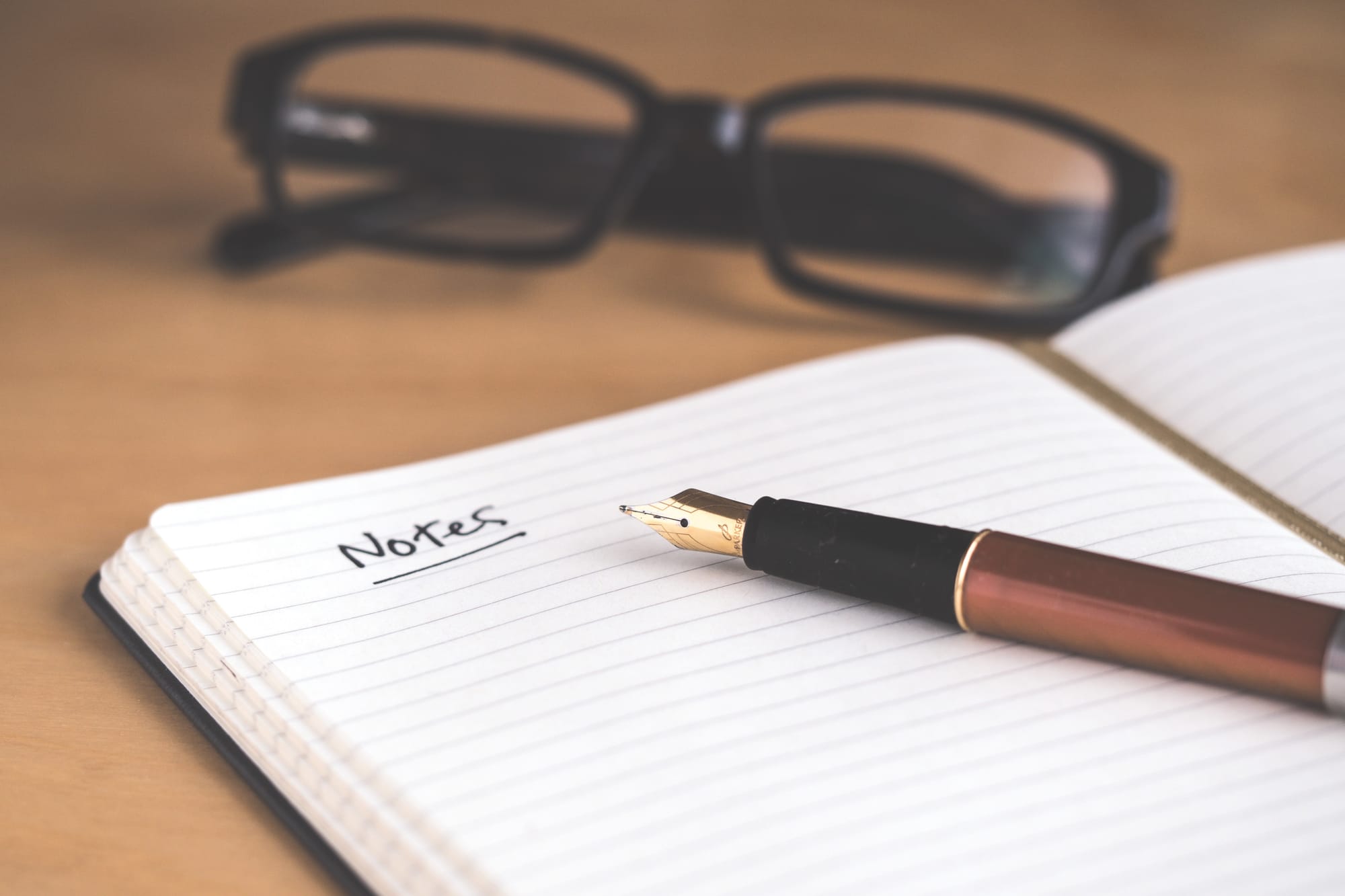 Notebook with fountain pen featuring the word "notes," underlined once. A pair of glasses sits above the notebook.